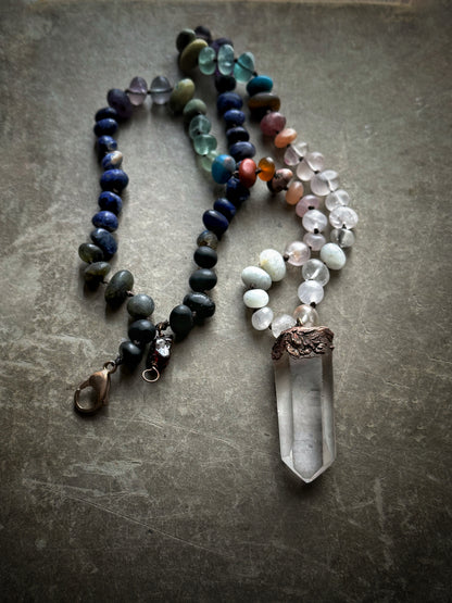 Collana annodata con pietre naturali miste effetto matte e ciondolo a punta grezza di quarzo ialino in rame, distesa su fondo grigio – Ebbijoux