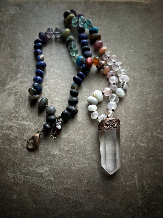 Collana annodata con pietre naturali miste effetto matte e ciondolo a punta grezza di quarzo ialino in rame, distesa su fondo grigio – Ebbijoux